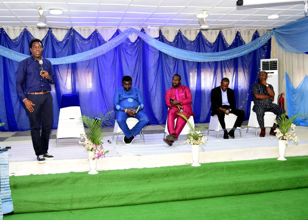 The host speaking to the room while four contributors sit on the stage